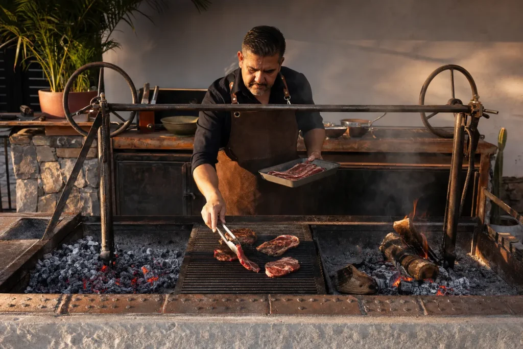 Asador comparando carbón o leña en una parrilla con brasas separadas y carne al centro.